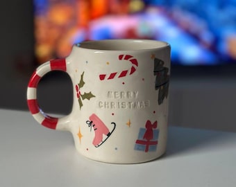 May include: Cream-colored ceramic mug with a red and white striped handle. The mug is decorated with Christmas-themed illustrations, including a candy cane, ice skate, and gift. The words "MERRY CHRISTMAS" are embossed on the front.