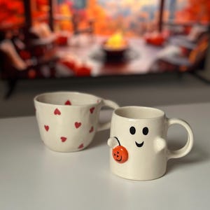 May include: Two ceramic mugs. One is off-white with red heart shapes. The other is a ghost-shaped mug with a smiling face and a small orange pumpkin. Both mugs have handles and are on a white surface.