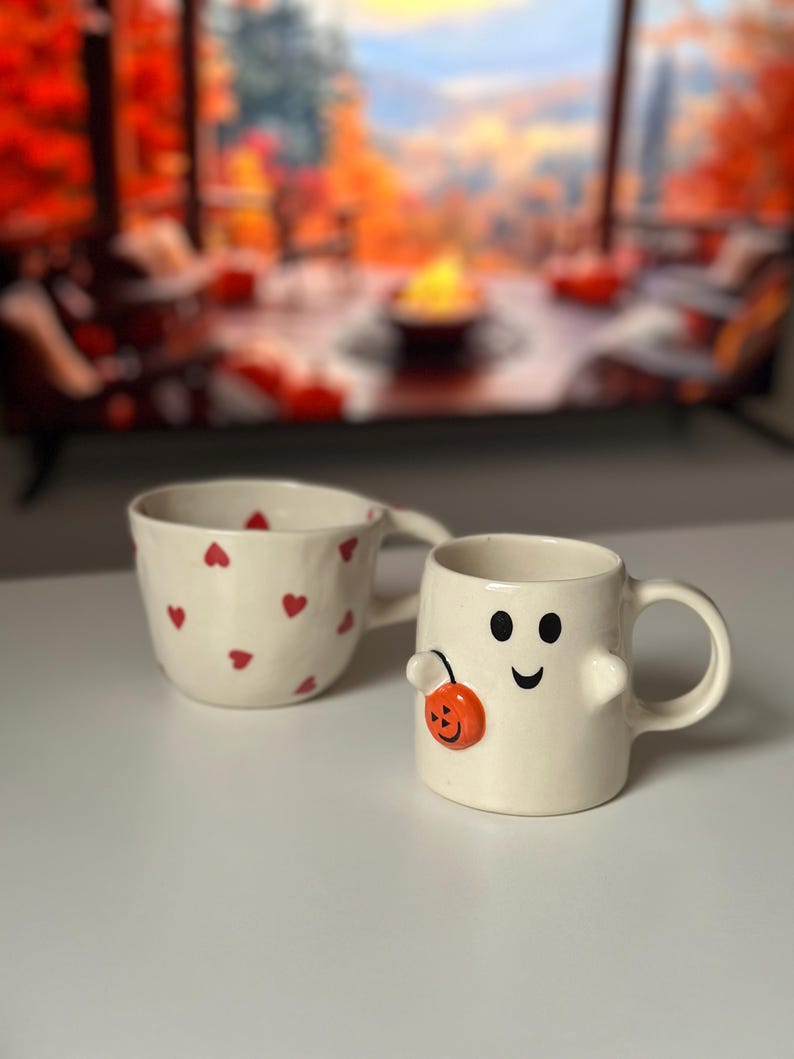May include: Two ceramic mugs. One is off-white with red heart shapes. The other is a ghost-shaped mug with a smiling face and a small orange pumpkin. Both mugs have handles and are on a white surface.