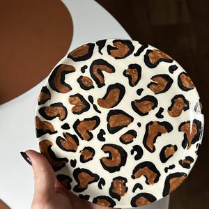 以下が含まれることがあります： ヒョウ柄のデザインが施された、丸い白いセラミックプレート。茶色と黒の斑点があり、斑点模様を作り出しています。白い背景と茶色の背景の中で、手でプレートを持っています。