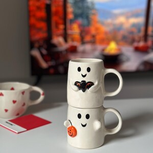 May include: Two stacked white ceramic mugs with ghost faces. The top mug has a black bat design, and the bottom mug has an orange pumpkin. A third mug with red hearts is visible in the background. Halloween-themed drinkware.