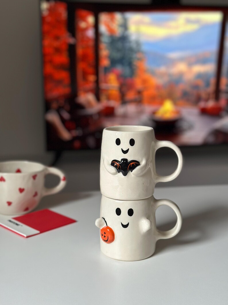 May include: Two stacked white ceramic mugs with ghost faces. The top mug has a black bat design, and the bottom mug has an orange pumpkin. A third mug with red hearts is visible in the background. Halloween-themed drinkware.