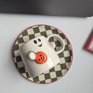 May include: A white ceramic Halloween-themed mug shaped like a ghost, with a smiling face and holding an orange pumpkin. The mug sits on a matching saucer with a green and white checkered pattern and a red rim. A festive seasonal item.