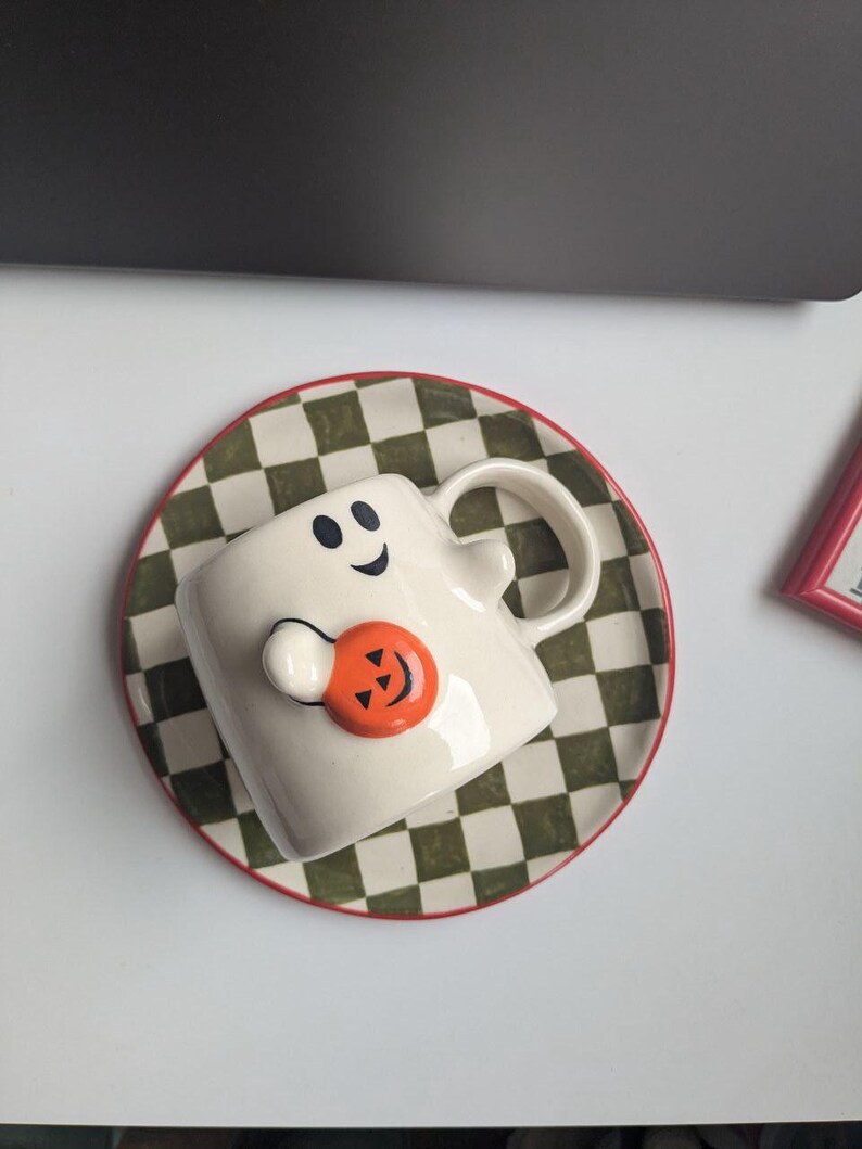May include: A white ceramic Halloween-themed mug shaped like a ghost, with a smiling face and holding an orange pumpkin. The mug sits on a matching saucer with a green and white checkered pattern and a red rim. A festive seasonal item.