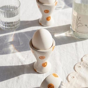 Peut inclure: Des coquetiers en céramique blanche ornés de visages souriants oranges, contenant chacun un œuf blanc. Les coquetiers sont posés sur une nappe blanche. Un verre d'eau et une bouteille sont en arrière-plan. Un ajout joyeux au petit-déjeuner.