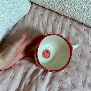 May include: A ceramic bowl with a red rim and a white interior. The exterior features a red and white checkered pattern. Inside the bowl is a pink smiley face with heart eyes. The bowl has a small handle.