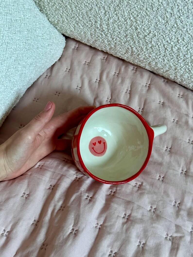May include: A ceramic bowl with a red rim and a white interior. The exterior features a red and white checkered pattern. Inside the bowl is a pink smiley face with heart eyes. The bowl has a small handle.
