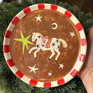 May include: A ceramic plate with a brown centre, decorated with a white horse with black star details, a pink bow, and a green star. The rim is white with red rectangles. The plate is a decorative item.