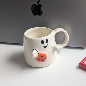 May include: A white ceramic Halloween mug shaped like a ghost, with a black smiling face and holding an orange pumpkin. The mug has a handle and is designed for beverages. A fun and festive item for the spooky season.