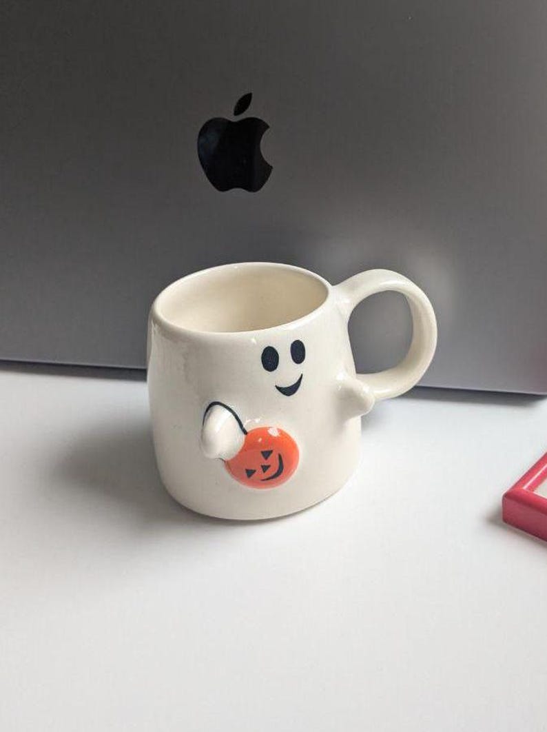 May include: A white ceramic Halloween mug shaped like a ghost, with a black smiling face and holding an orange pumpkin. The mug has a handle and is designed for beverages. A fun and festive item for the spooky season.