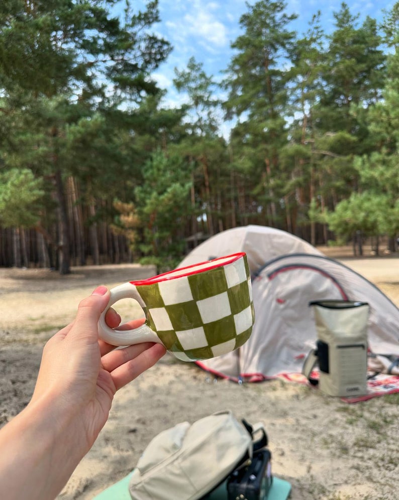 May include: A ceramic mug with a white handle and a green and white checkered pattern. The rim is red. The mug is held in front of a tent and trees.