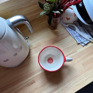 May include: A white ceramic mug with a red rim and a small handle. The inside of the mug features a smiley face design. A white electric kettle and a small vase of flowers are also visible on a wooden surface.