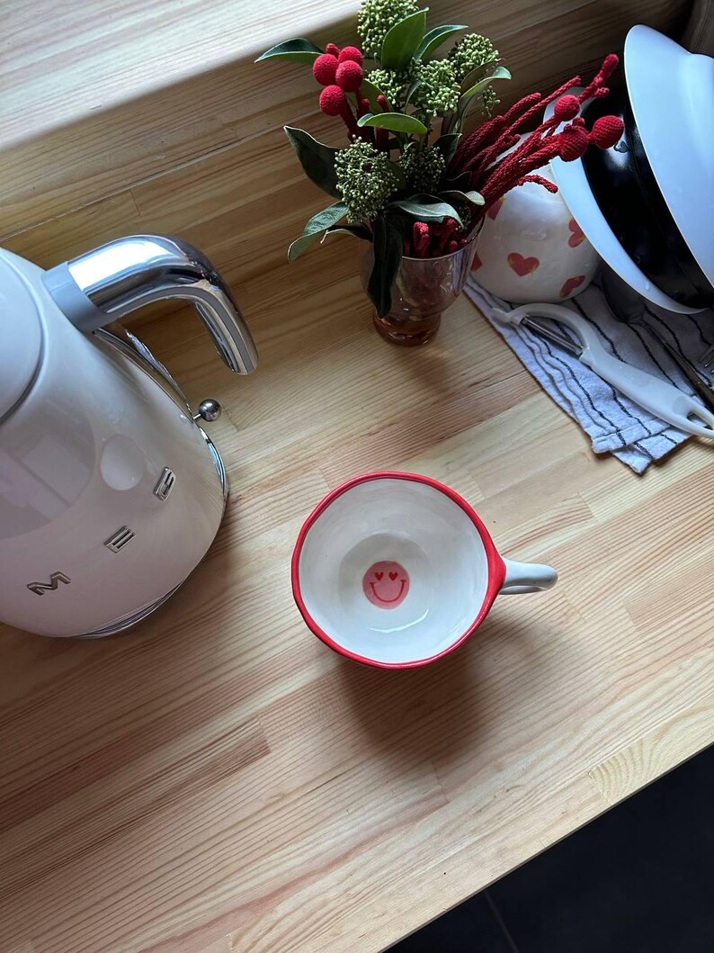 May include: A white ceramic mug with a red rim and a small handle. The inside of the mug features a smiley face design. A white electric kettle and a small vase of flowers are also visible on a wooden surface.