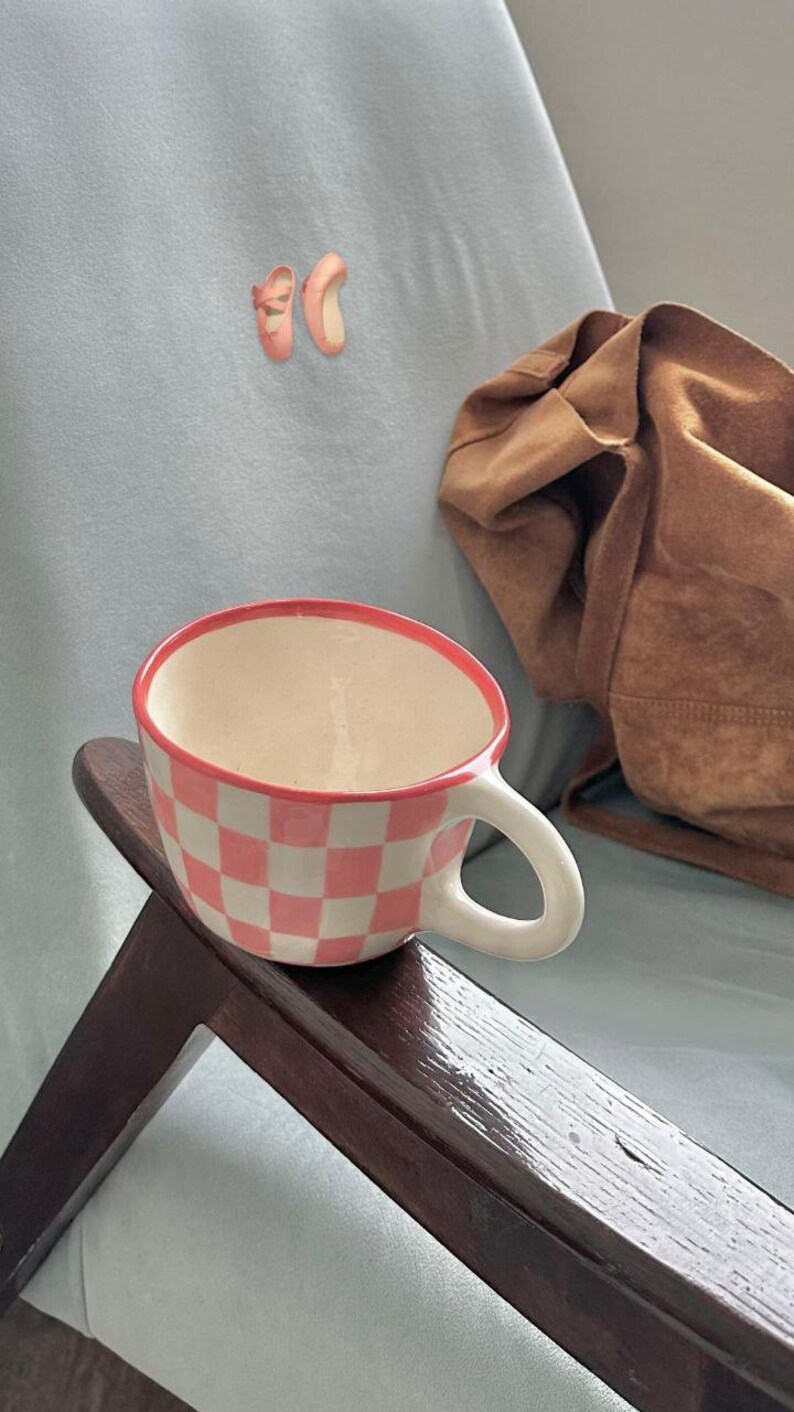 May include: A ceramic coffee mug with a pink and white checkered pattern. The rim is outlined in pink. The mug sits on a dark wooden surface. A pair of pink ballet slippers are pinned to the light blue background.