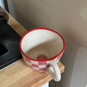 May include: A ceramic mug with a red and white checkered pattern and a red rim. The mug has a white handle and a small smiley face at the bottom. The mug is sitting on a wooden surface.