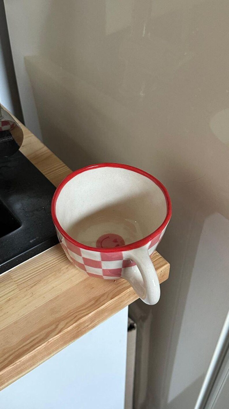 May include: A ceramic mug with a red and white checkered pattern and a red rim. The mug has a white handle and a small smiley face at the bottom. The mug is sitting on a wooden surface.