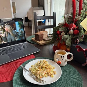 May include: A laptop displaying a video, a plate of food with a fork, and a mug with a snowman face on a table. A Christmas decoration with red candles and ornaments is in the background. Slices of bread are on a wooden board.