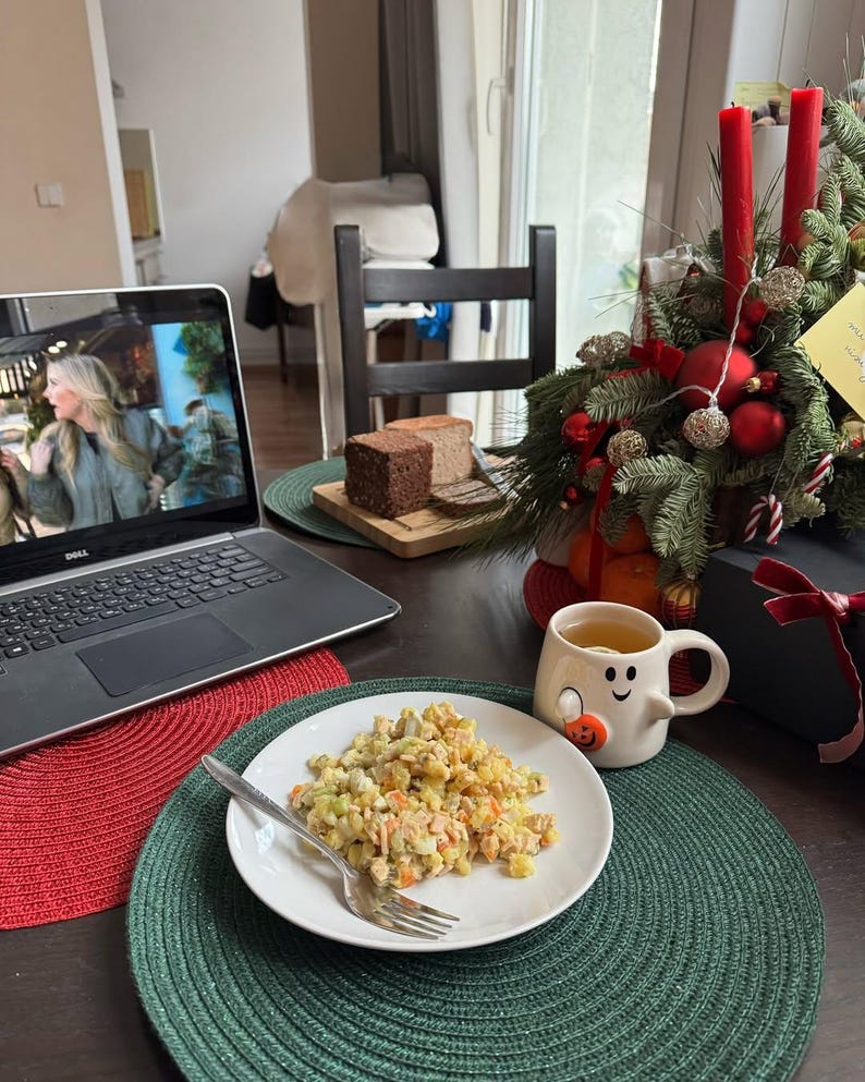 May include: A laptop displaying a video, a plate of food with a fork, and a mug with a snowman face on a table. A Christmas decoration with red candles and ornaments is in the background. Slices of bread are on a wooden board.