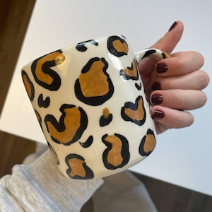 May include: A ceramic mug with a cream-colored base and a leopard print pattern in brown and black. The mug has a curved handle and a square shape.