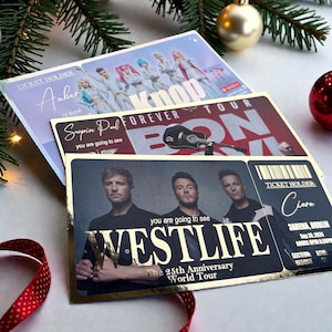 May include: Three concert ticket holders with gold foil detailing are displayed. One features a K-pop group, another a guitar, and the third, the band Westlife. The Westlife ticket is for the 25th Anniversary World Tour.