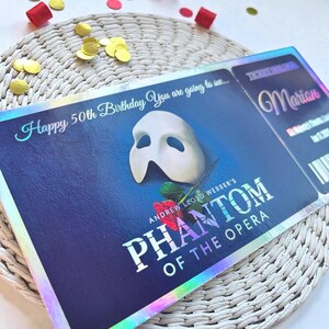 May include: A Phantom of the Opera ticket with a white mask and red rose on a dark blue background. The ticket is for Marion's 50th birthday. Gold confetti and red ribbon are scattered around the ticket.