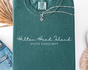 May include: A dark teal crewneck t-shirt featuring the white text Hilton Head Island and SOUTH CAROLINA with a small palm tree icon. The shirt has a Comfort Colors label at the inner neckline and is displayed with a silver chain necklace.