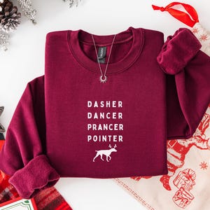 May include: A maroon long-sleeve crewneck sweatshirt featuring white text that reads DASHER, DANCER, PRANCER, POINTER, followed by a white graphic of a dog with antlers. A silver sunburst pendant necklace rests on the neckline of the garment.
