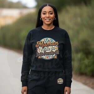 Puede incluir: Sudadera negra y pantalón a juego con el texto "Maman reine du je te l'avais dit" en un diseño circular. El diseño incluye colores dorados, naranjas, azules y rojos. La mujer lleva pendientes de aro dorados.