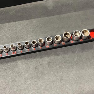 Può includere: Un organizzatore di bussole in plastica nera con 15 slot per diverse dimensioni di bussole. Le bussole sono argentate e i numeri 4, 4,5, 5, 5,5, 6, 7, 8, 9, 10, 11, 12, 13, 14 e 15 sono stampati in rosso sull'organizzatore.