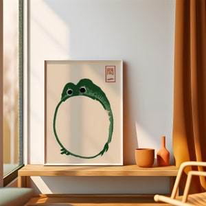 Puede incluir: Una impresión enmarcada de una rana verde en un círculo con el carácter japonés "間" en rojo sobre un fondo blanco. La impresión está colgada en una pared sobre un estante de madera con una maceta marrón y un jarrón.