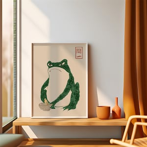 May include: A framed print of a green frog holding a bowl and a wooden stick. The frog is sitting on a beige background with the Japanese character "間" in red above it.