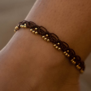 Peut inclure: Un bracelet de cheville en macramé marron avec des perles dorées. Le bracelet est porté sur une cheville, mettant en valeur le design tissé complexe et le contraste entre le cordon marron foncé et les perles brillantes. Un bijou simple et élégant.