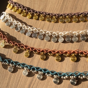 May include: Close-up of several macrame necklaces in various colors, including brown, white, rust, and teal. Each necklace features spiral-shaped metal charms and small beads. The necklaces are displayed on a wooden surface.