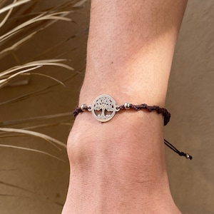 Pode incluir: Uma pulseira com um berloque prateado de árvore da vida e uma pulseira tecida marrom escuro. A pulseira apresenta um berloque circular com um intrincado design de árvore e pequenas contas metálicas. A pulseira é ajustável.