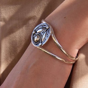 May include: Silver cuff bracelet featuring an oval centerpiece with a detailed design. The centerpiece has a central stone and is surrounded by decorative elements. The bracelet is worn on a wrist, showcasing its elegant design and craftsmanship.
