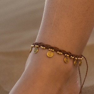 May include: Brown woven anklet with gold-colored spiral charms and small beads. The anklet is worn on an ankle, with the background out of focus.
