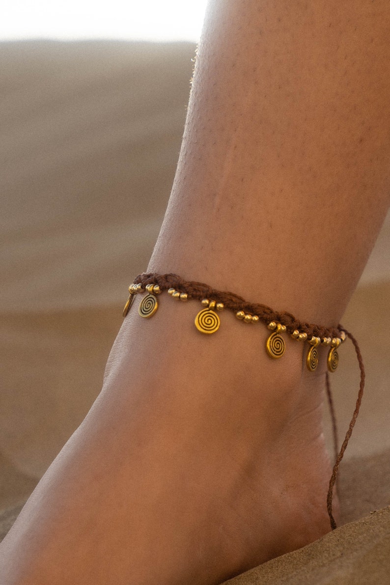 May include: Brown woven anklet with gold-colored spiral charms and small beads. The anklet is worn on an ankle, with the background out of focus.