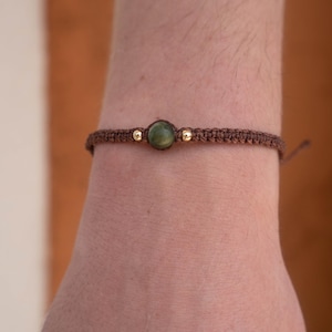 Può includere: Un braccialetto in macramè marrone con una pietra verde centrale e due piccole perline dorate. Il braccialetto è annodato e sembra regolabile. Il braccialetto è indossato su un polso.