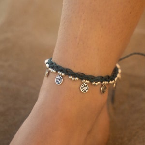 May include: Dark teal anklet with silver-colored spiral charms and small beads. The anklet is worn on an ankle, showcasing the design and details of the jewelry. The background is a blurred, neutral tone.