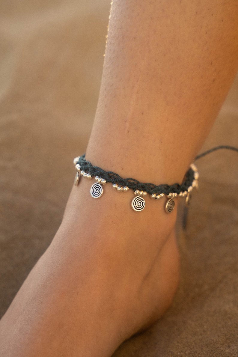 May include: Dark teal anklet with silver-colored spiral charms and small beads. The anklet is worn on an ankle, showcasing the design and details of the jewelry. The background is a blurred, neutral tone.
