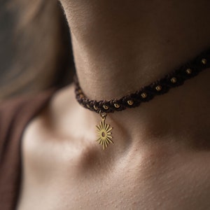 Peut inclure: Un collier ras de cou tressé marron avec un pendentif soleil en or. Le collier est porté par une personne au teint clair.