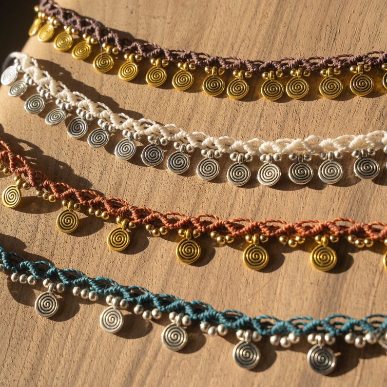 May include: Close-up of several macrame necklaces in various colors, including brown, white, rust, and teal. Each necklace features spiral-shaped metal charms and small beads. The necklaces are displayed on a wooden surface.