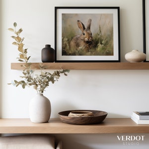 May include: A framed painting of a brown rabbit sitting in a field of green grass. The painting is hanging on a white wall above a wooden shelf with a vase of dried leaves and a wooden bowl.