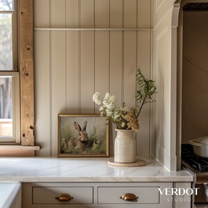 May include: A white ceramic vase containing a bouquet of white flowers and dried greenery sits on a white countertop. A framed print of a rabbit in a field is on the counter behind the vase. The countertop is in front of a wall with vertical wood paneling.