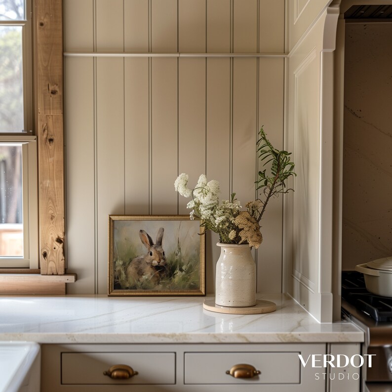 May include: A white ceramic vase containing a bouquet of white flowers and dried greenery sits on a white countertop. A framed print of a rabbit in a field is on the counter behind the vase. The countertop is in front of a wall with vertical wood paneling.