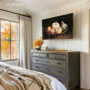 May include: A bedroom scene featuring a dark gray dresser with four drawers and a light wood top. A large framed artwork of pumpkins hangs above the dresser. A window with fall foliage is visible.