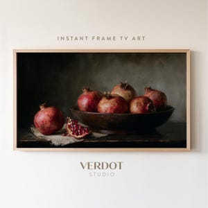 Peut inclure: Peinture encadrée de nature morte avec des grenades. L'œuvre représente plusieurs grenades entières et une grenade coupée, disposées dans un bol en bois et sur un tissu. Le texte "INSTANT FRAME TV ART" et "VERDOT STUDIO" sont visibles.