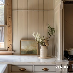 May include: A white ceramic vase with a bouquet of dried white flowers and greenery sits on a wooden coaster on a white marble countertop. The countertop is in front of a wall with vertical wood paneling painted a light beige color.