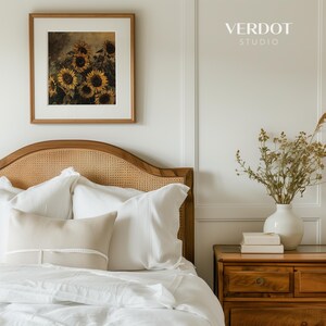 May include: A white bed with a wicker headboard and white bedding. There are two white pillows and a beige pillow with white trim on the bed. A framed picture of sunflowers hangs on the wall above the bed. A wooden nightstand with a vase of dried flowers and books sits to the right of the bed. The text "VERDOT STUDIO" is printed on the wall above the nightstand.