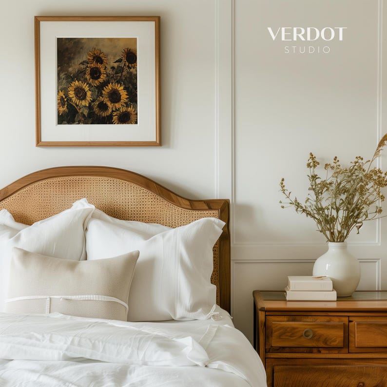 May include: A white bed with a wicker headboard and white bedding. There are two white pillows and a beige pillow with white trim on the bed. A framed picture of sunflowers hangs on the wall above the bed. A wooden nightstand with a vase of dried flowers and books sits to the right of the bed. The text "VERDOT STUDIO" is printed on the wall above the nightstand.